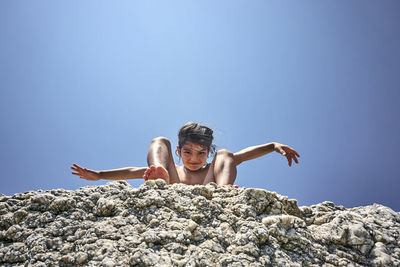 Low angle portrait of shirtless girl on rocks against clear blue sky during sunny day