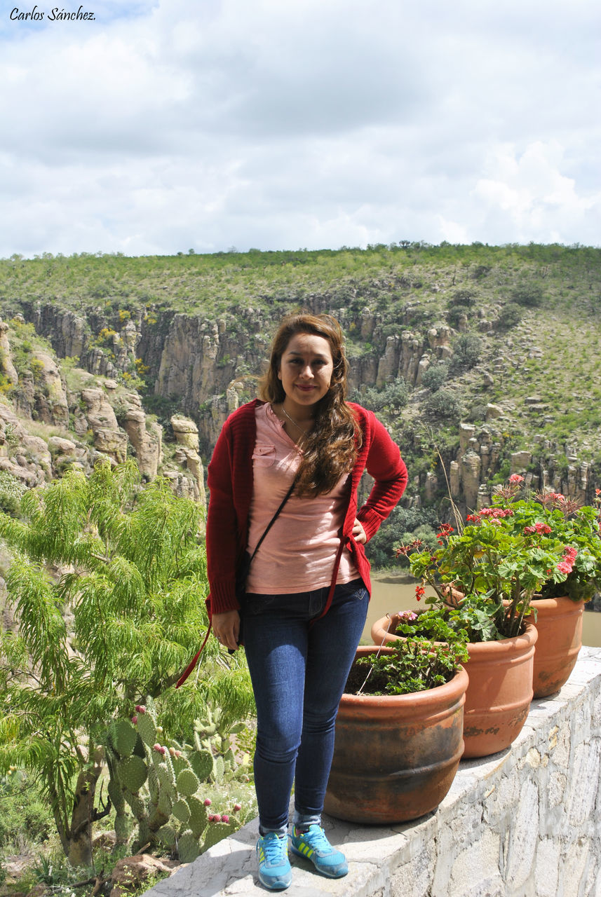 PORTRAIT OF YOUNG WOMAN STANDING ON ROCK AGAINST SKY