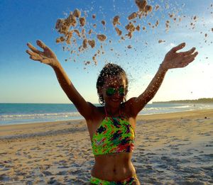 Happy young woman throwing sand at beach against sky