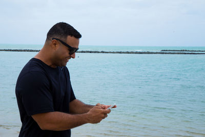 Young man using mobile phone at beach against sky