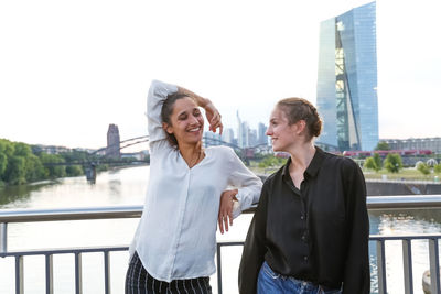 Cheerful young women standing against railing on bridge in city