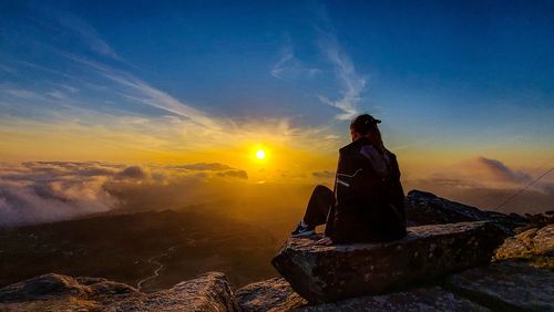 Woman sitting on rock against sky during sunset