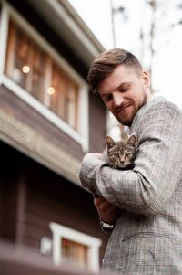 Low angle view of handsome young man carrying cat while standing against building in city