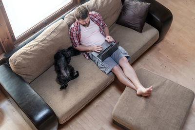 Man works with laptop, he sitting on sofa, next to his dog schnauzer sleeping with stomach upstairs