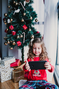 Portrait of beautiful kid girl at home by the christmas tree and using mobile phone