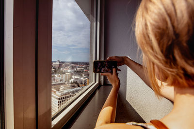 Rear view of woman taking selfie by window at home