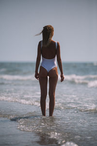 Rear view of seductive woman wearing one piece swimsuit while standing on shore at beach