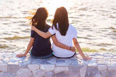 Rear view of friends looking at sea while sitting on retaining wall during sunset