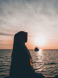Woman wearing hijab looking away while sitting by sea against sky during sunset