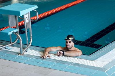 Bearded male athlete in swimming cap and goggles watching time on wristwatch while leaning on poolside