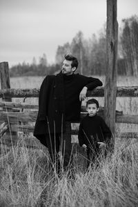 Father and son in black clothes stand in a field with an old wooden horse fence in the fall