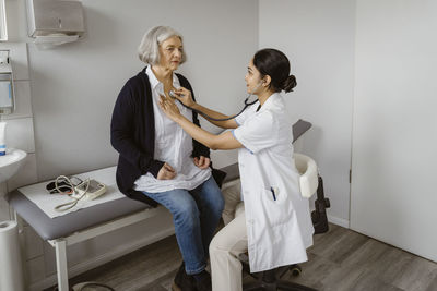 Side view of female doctor listening heartbeat of senior patient sitting on bed in clinic