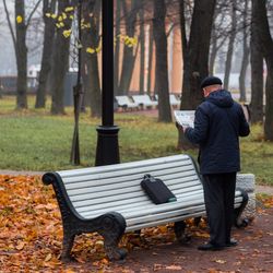 Full length rear view of man reading newspaper by bench in park during autumn