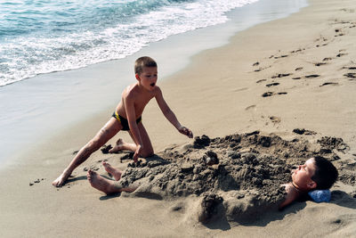 Boy looking at brother buried in sand on beach