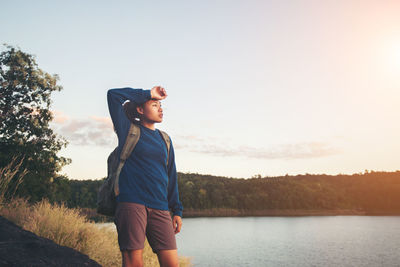 Tired female hiker carrying backpack while standing at lakeshore against clear sky