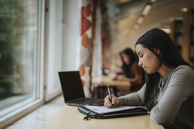 Side view of student writing in paper while studying in library