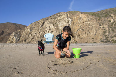 Girl playing with sand while sitting by dog at beach against mountains