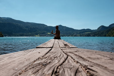 Mid adult man with beer bottle sitting on pier over lake against clear blue sky