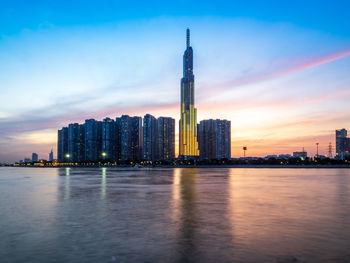 River by illuminated buildings against sky during sunset