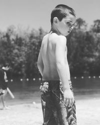 Shirtless boy standing at lakeshore during sunny day