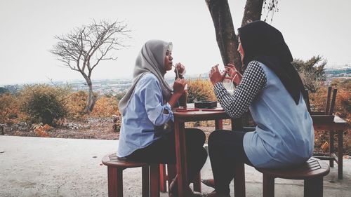 Side view of female friends talking while sitting at outside cafe against sky