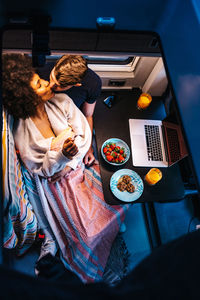 From above of happy multiracial couple kissing gently while having meal near laptop as resting together in camper van during vacation