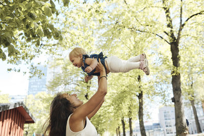 Side view of playful mother lifting daughter against trees