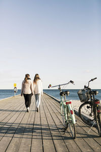 Rear view of female friends walking on pier with bicycles in foreground at beach