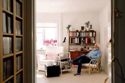 Full length of senior man sitting on chair in living room seen from doorway