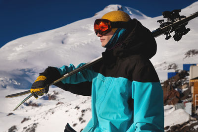 A portrait of a professional skier in an orange ski goggles stands with skis on his shoulder against