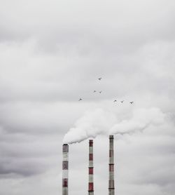 Birds flying over smoke emitting chimneys against cloudy sky