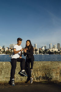 Full length of young couple using smart phone while exercising on promenade against cityscape