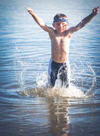 Shirtless boy wearing swimming goggles with arms raised standing in lake