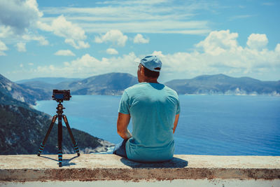 Rear view of man sitting by camera on retaining wall while looking at sea against sky