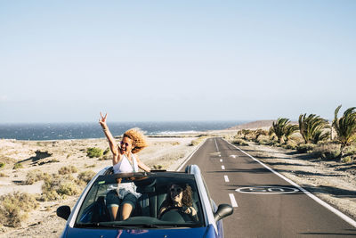 Woman blowing a kiss while peeking out from with friend driving car against sky