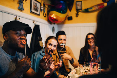 Young friends holding props while sitting at restaurant during dinner party