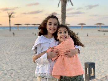 Portrait of smiling two young girls hugging  standing against sky near the beach. 