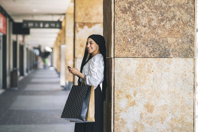 Smiling young woman with mobile phone looking away leaning on column