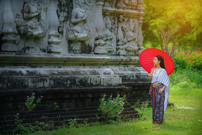 Woman with red umbrella standing by temple on grassy field