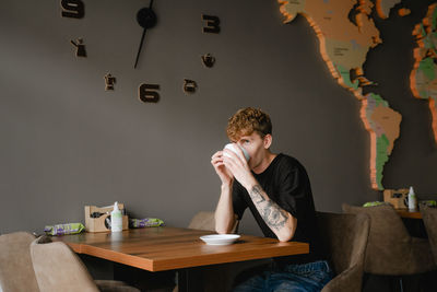 A guy with a tattoo on his arm sits at a table in a public place, drinks coffee from a white glass