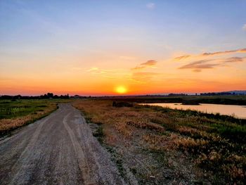 Dirt road amidst field against sky during sunset