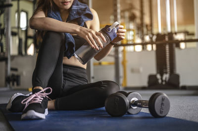 Low section of woman holding water bottle sitting on exercise mat in gym