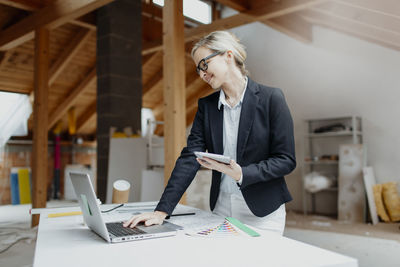 Mid adult woman holding digital tablet while using laptop on table in workshop