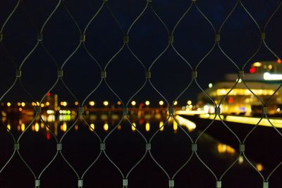 Close-up of chainlink fence against illuminated city at night