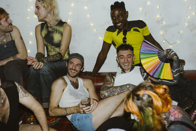 Happy non-binary friends sitting on sofa at enjoying party at illuminated nightclub