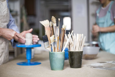 Hand tools and paintbrushes in containers on workbench with female potters working in background