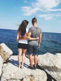 Rear view of couple with arms around standing on cliff by sea
