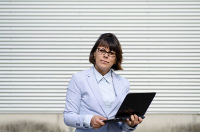 Portrait of businesswoman holding laptop against shutter