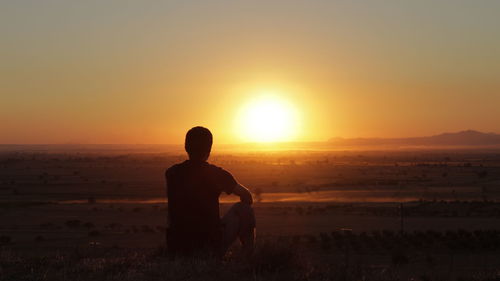 Rear view of man sitting on field against sky during sunset