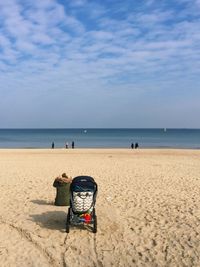 Rear view of woman with baby stroller sitting at beach against sky